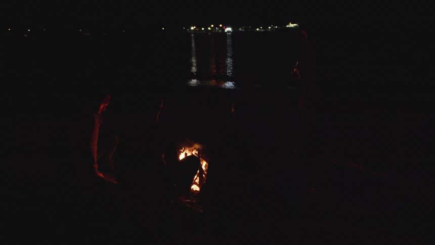 A fire burns on the ground people sit nearby water reflects lights in the distance a dark night surrounds the scene