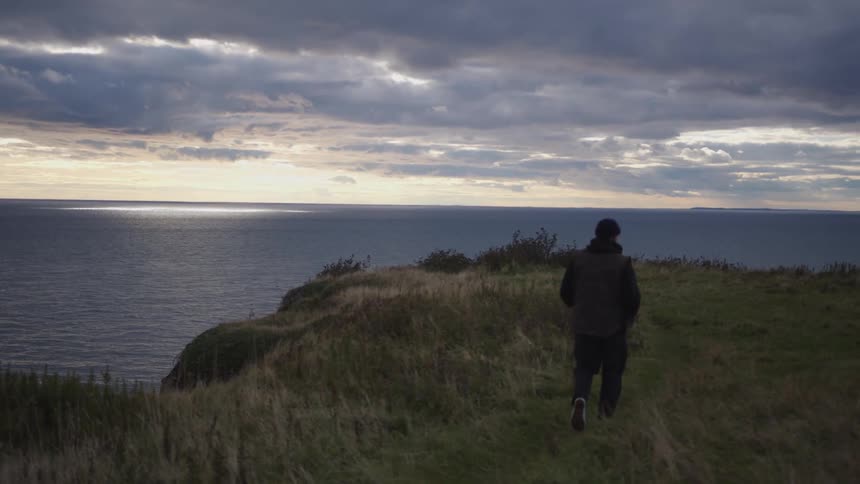 Man walks along a grassy cliff overlooking the sea at sunset