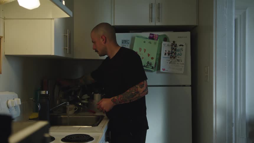 A young man with a tattooed arm does the dishes at the kitchen sink while wearing a black tshirt
