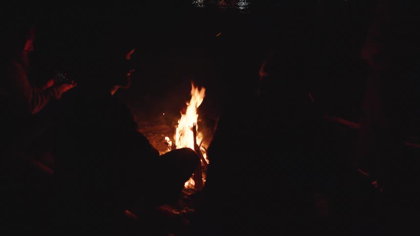Three men sitting by a bonfire next to a body of water talking outdoors during night time
