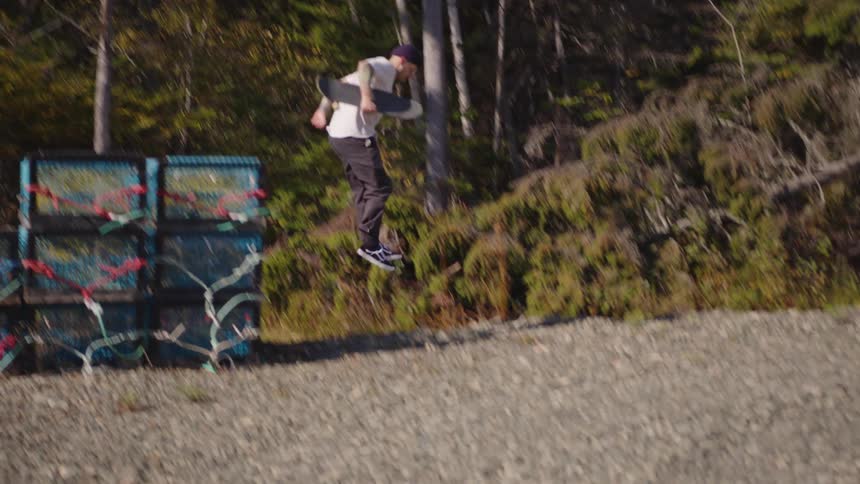 A man carrying a skateboard steps off a stack of crates and jumps down to the gravel ground landing with a crouching motion in front of a dense line of trees