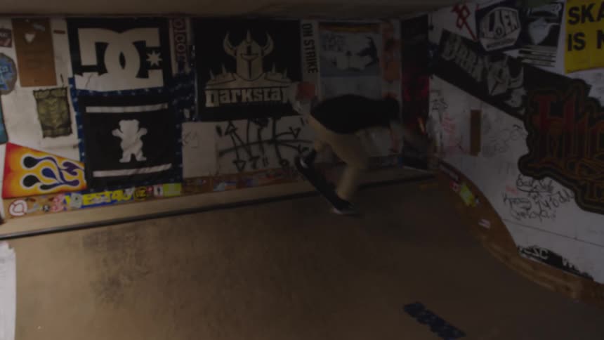The video shows a person skateboarding inside a dimly lit indoor space possibly a homebuilt skate park that is decorated with posters and graffiti the skater is practicing on a small wooden quarter pipe or ramp watched by another person