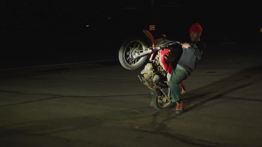 Motorcyclist performs wheelie on red bike on paved area at night in front of lit building