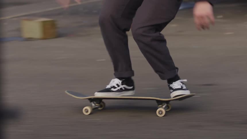 A close moving view focuses on the legs of a person wearing black pants and black and white vans sneakers riding a skateboard down a dark paved road