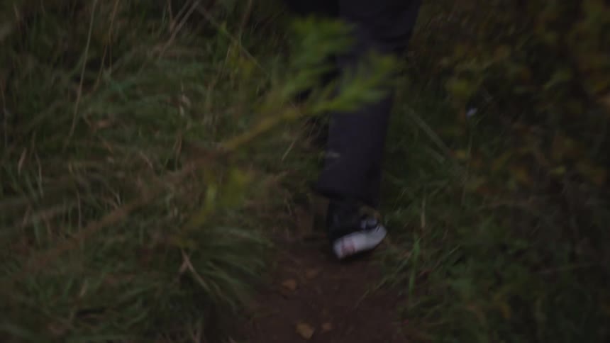 The video shows a lowangle closeup shot of a persons legs walking over rough uneven terrain covered in dark green grass and brush the person is wearing dark pants and white and black athletic shoes while moving through the outdoor environmen
