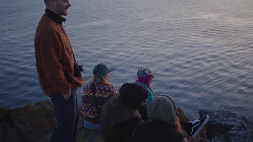 Friends gather on a rocky cliff overlooking water at sunset