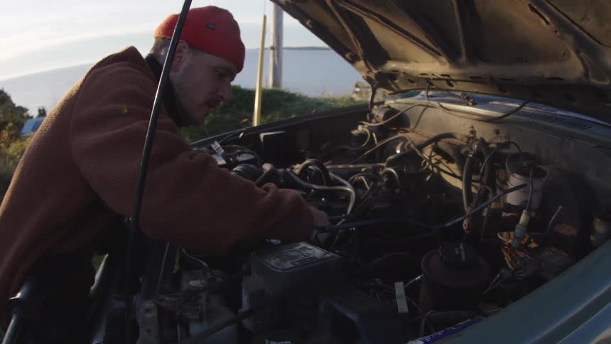 Man in orange beanie performs maintenance under vehicle hood in outdoor setting near water