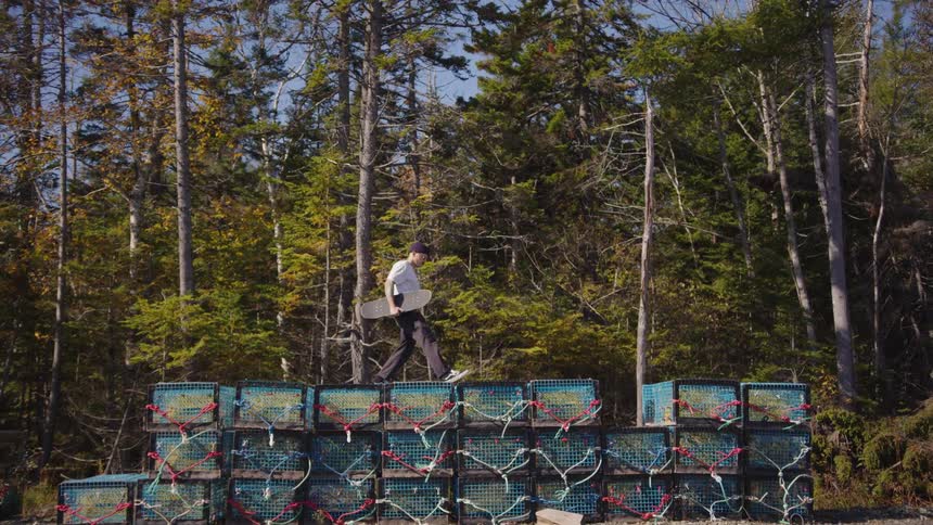 A young man carrying a skateboard walks across a tall layered stack of blue and black lobster traps against a backdrop of dense forest under a bright sky