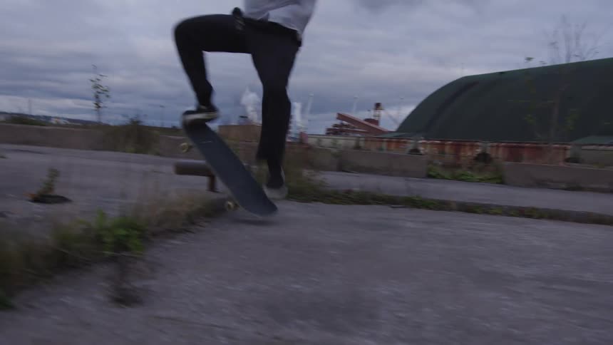 A man wearing a white t shirt dark pants and a yellow beanie skateboards on a gray concrete slab in an abandoned industrial area with large covered buildings and smoking chimneys in the background under an overcast sky