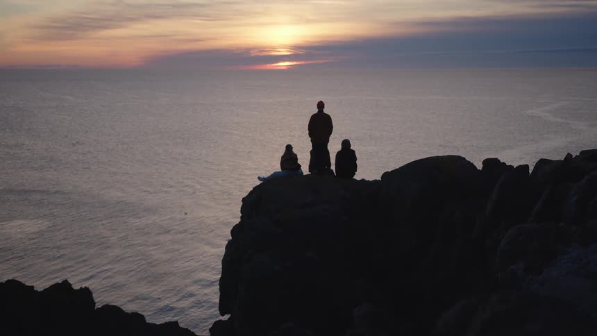 The video is a wide establishing shot of three people standing on a silhouetted rocky cliff overlooking the vast ocean at sunset they are standing close together dwarfed by the landscape enjoying the view of the sun setting over the water which