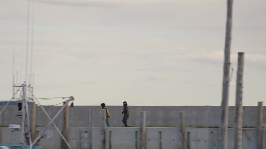 Two men in dark casual clothing walk along a wide concrete seawall at a harbor or port on an overcast day with a fishing boat and masts visible in the foreground
