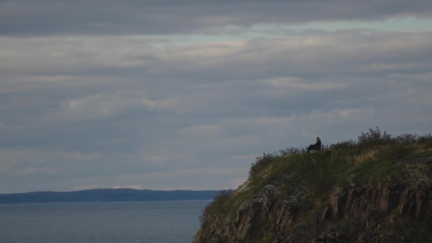 A lone figure sits on the steep edge of a rocky cliff covered with low brush overlooking a large expanse of gray water and a distant shore under a wide cloudy sky
