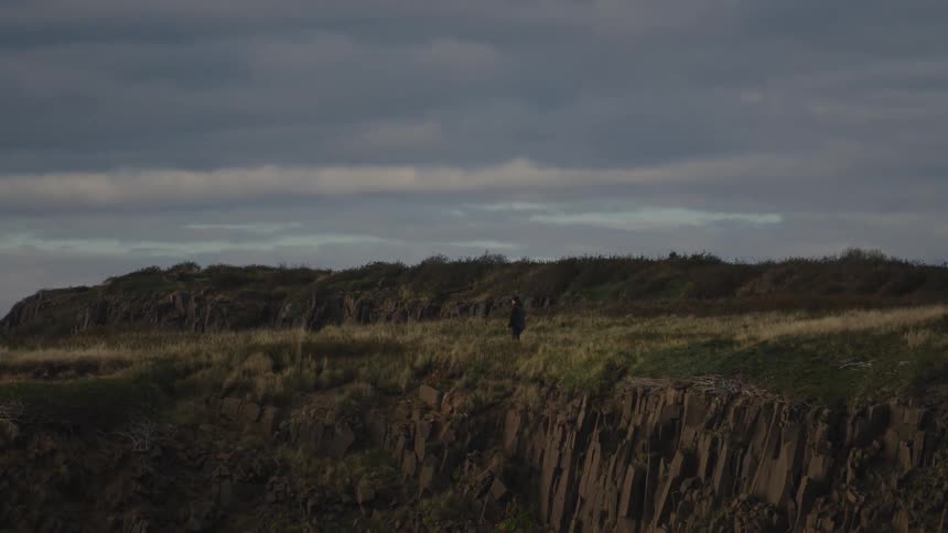 Person walks alone on a grassy cliff under a cloudy sky