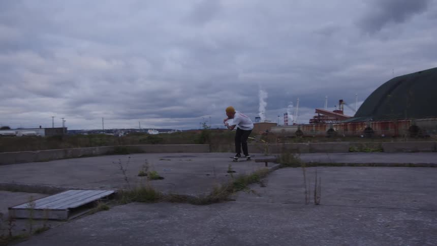 A man wearing a white shirt and yellow beanie skateboards on a flat concrete slab in a large abandoned industrial area with factories and a covered building in the background under an overcast sky