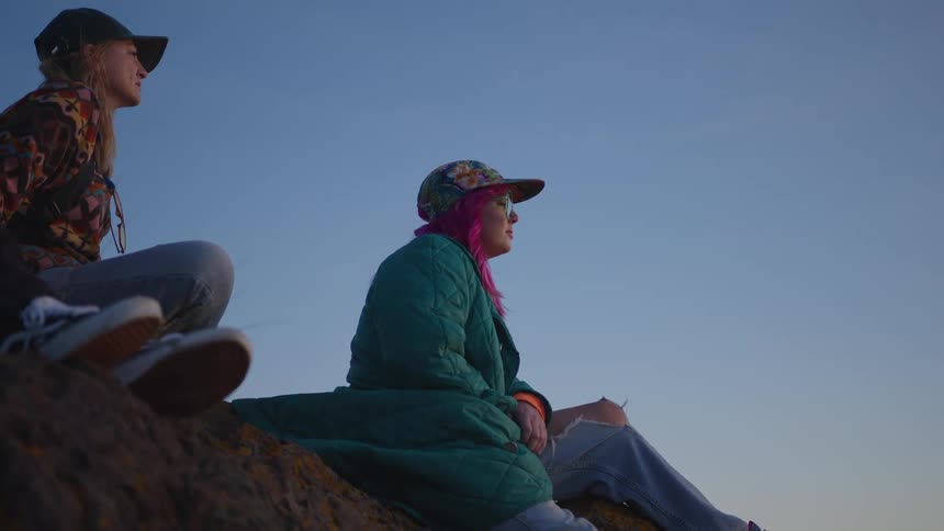 Three white women sitting down on a rocky slope outdoors during dusk or dawn