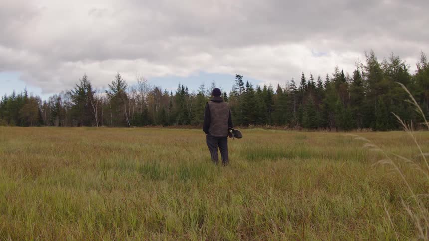 Person standing in an open field holds a skateboard while looking at the forest