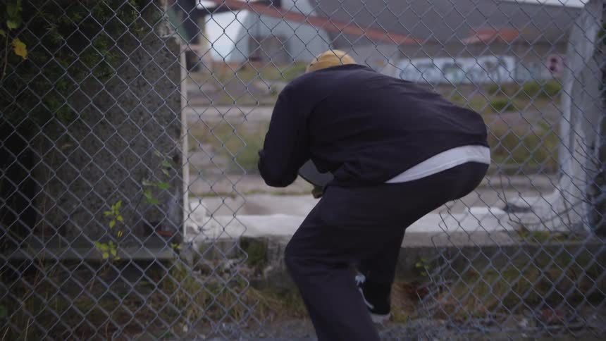 A person wearing dark clothes and a cap holds a skateboard ducks under a metal fence steps up onto concrete and moves away from the camera toward structures