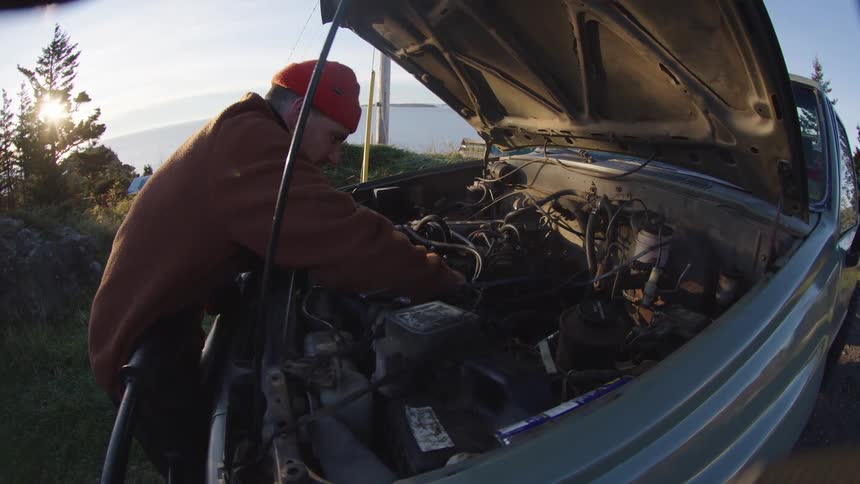 The video is a wideangle shot of a person wearing an orange beanie and brown jacket working on the engine of a vehicle outdoors the hood is fully open and they are bent over actively inspecting or performing maintenance on the engine block