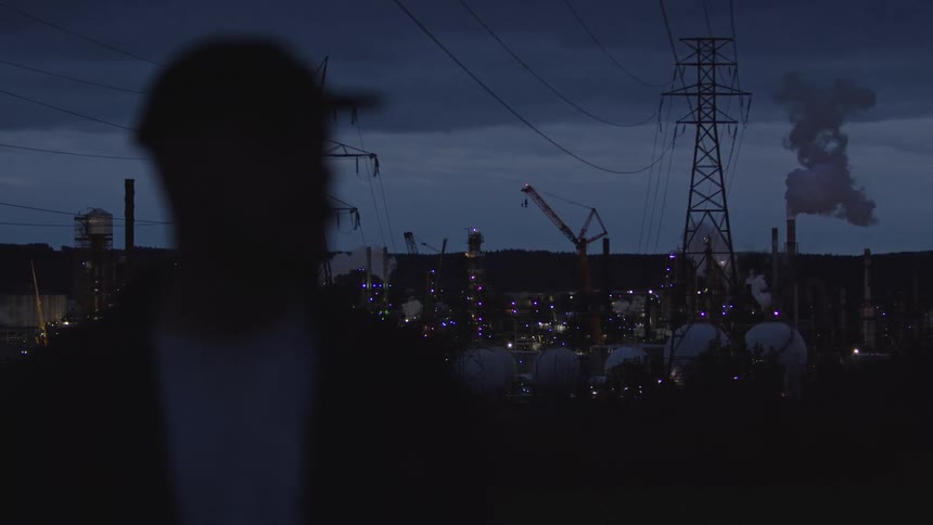 Silhouette of a man in the foreground with a lit refinery in the background at night