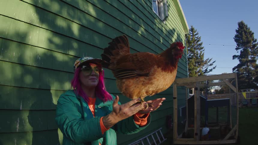 A woman holding a chicken on her palms near a house at sunset