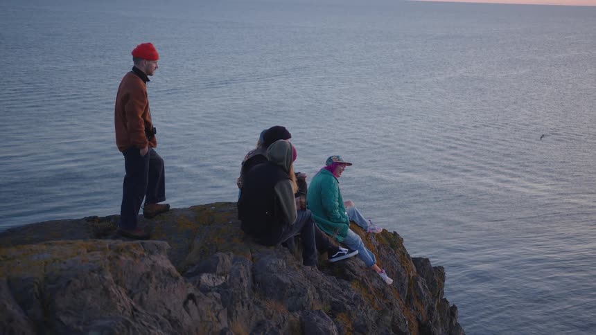 Man stands behind his friends as they sit on a promontory overlooking the sea during the daytime