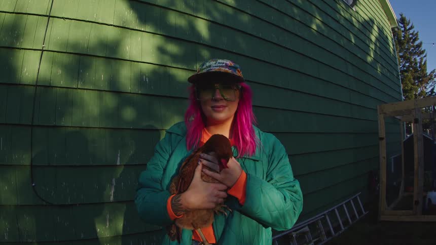Woman with pink hair holds a chicken in her arms beside a green wall under lateafternoon light