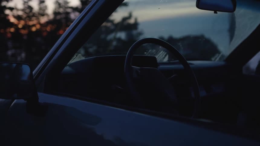 A car is parked on the side of the road at sunset its windows rolled down as golden light spills across the pavement and reflects softly off the vehicles surface