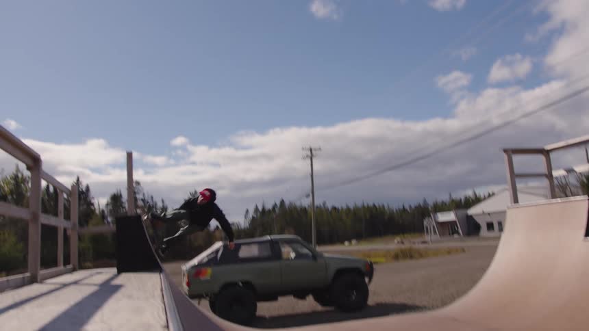 A skateboarder pushes off a ramp and performs a large jump over a parked truck landing the trick with another person sitting nearby and utility poles visible against a cloudy sky