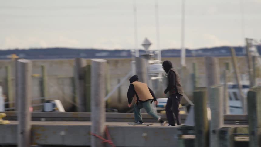 Two people wearing dark clothing and hoods run along a concrete pier with wooden pilings and docking equipment in the foreground past fishing boats and trucks on a dull overcast day