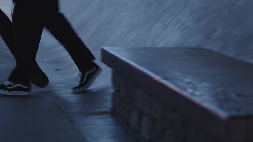 A low view focuses on the legs and feet of a person in black pants and vans sneakers stepping onto a skateboard and riding away past a concrete bench in a dimly lit skatepark