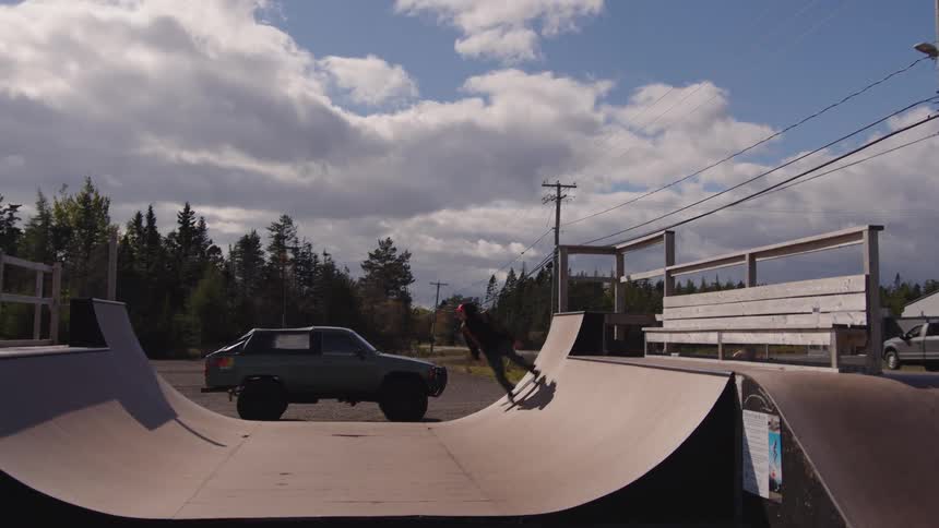 The video is a wide shot of a person skateboarding a halfpipe at an outdoor skate park a darkcolored pickup truck is parked in the middle of the ramps flat bottom forcing the skater to ride around it