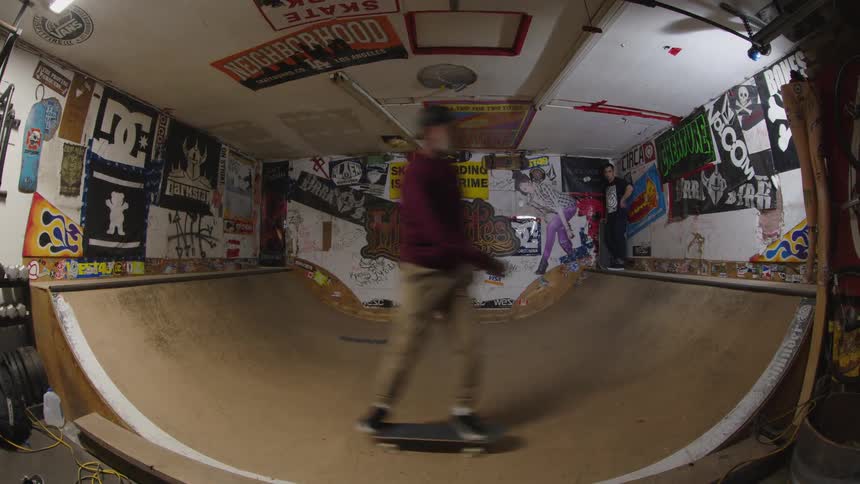 Skater performs trick on wooden halfpipe surrounded by graffiti posters and central slogan about skate culture