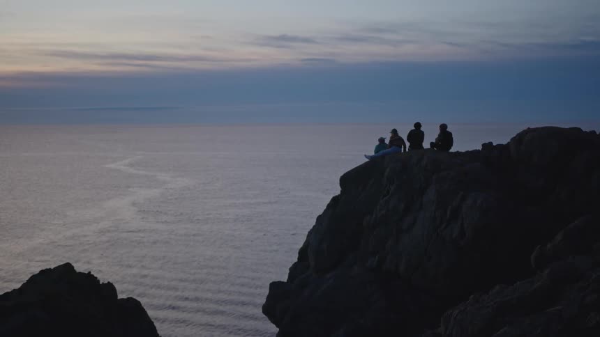Four friends sit on a cliff along the shoreline at sunset