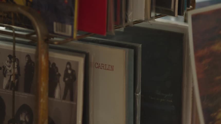 The video is a closeup shot inside a record store showing a persons hand with a tattoo browsing through a rack of vinyl record albums the records are tightly packed with one album cover prominently featuring the name carlin in the foreg