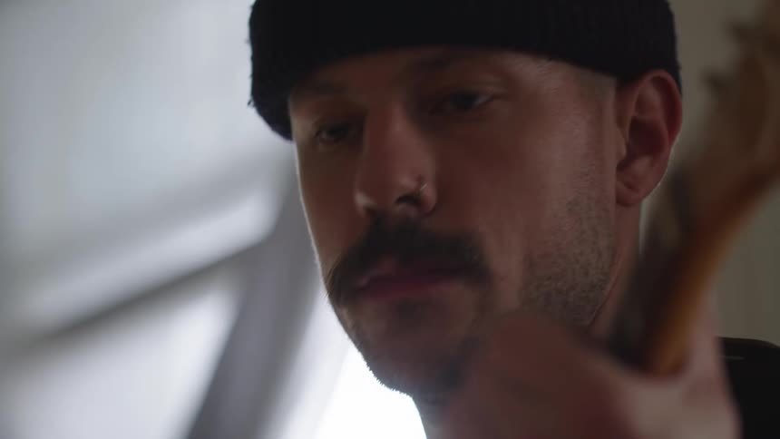 A man wearing a dark knit cap and a nose ring plays a guitar looking into the camera before turning his head to present his profile