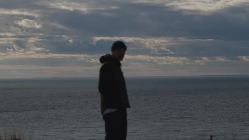 A man wearing a jacket stands on a cliff at sunset gazing out over the vast ocean as golden light bathes the horizon casting long shadows