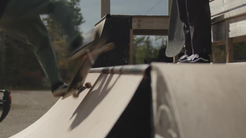 A person riding a skateboard quickly slides across the coping of a wooden halfpipe ramp in an outdoor skatepark with their feet and lower legs briefly visible above the ramps edge while other person wait in the top of the half pipe