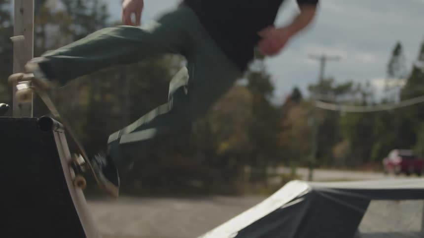 A skateboarder in dark clothing and a red cap performs a jump over the edge of a skate ramp in the foreground with a forested area and a road visible in the background