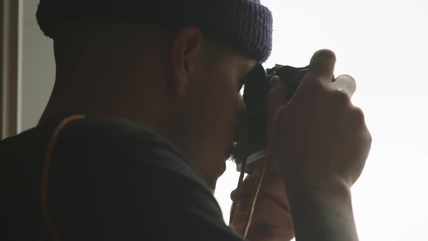 A man wearing a purple beanie and dark shirt stands in front of a bright window holding a camera up to his eye and then lowers the camera and looks out the window