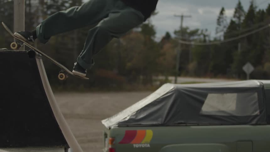 The video captures a person quickly skateboarding off a ramp into the air in the foreground in the background there is a green pickup truck with a black camper shell parked and the setting appears to be a rural or wooded area