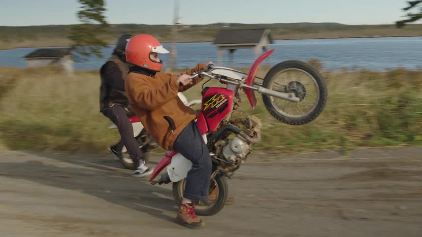 Two men wearing helmets perform a wheelie on a red motorcycle on a dirt road near a lake