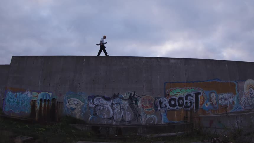 Person walks with a skateboard over a concrete wall covered in graffiti at dusk
