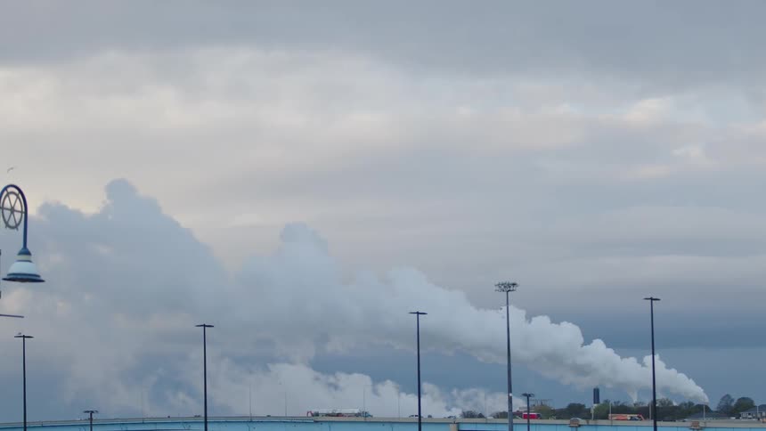 Industrial smoke rising into a cloudy sky above a bridge and city lights
