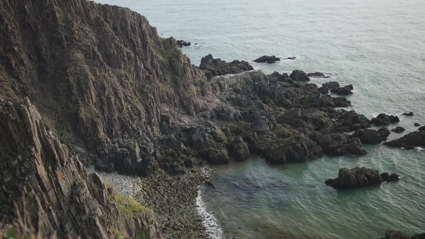 A camera captures a highangle view of a vast sheer layered rock cliff face descending to meet a rocky shoreline where greentinted ocean water laps against the dark stone