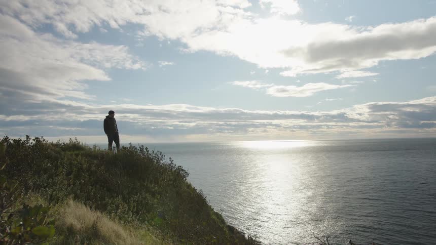 The video is a wide shot of a solitary person standing on a grassy cliff edge they are looking out over a vast ocean or sea with the sun reflecting brightly on the water beneath a partly cloudy sky