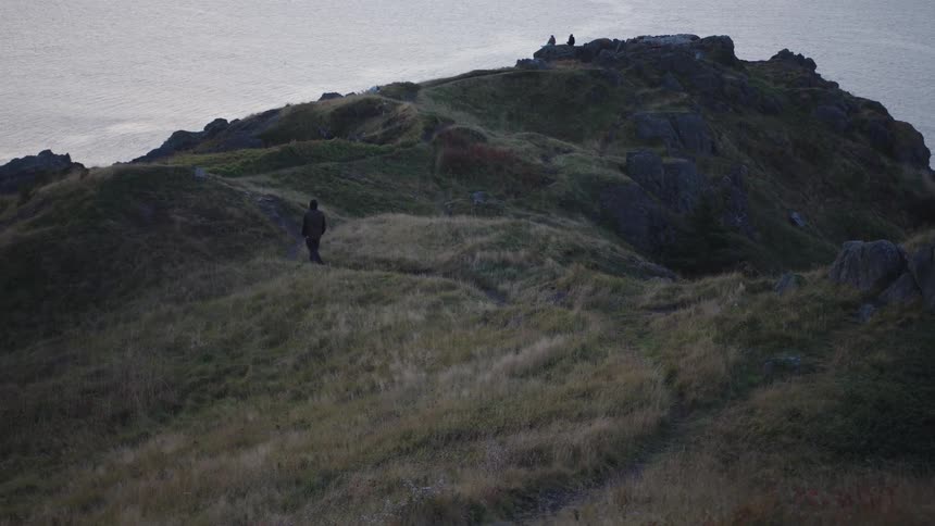 The video shows a lone person walking slowly down a grassy rugged hillside with patches of rock under a dark overcast sky the figure is silhouetted in a dark coat moving down the slope toward the foreground with the vast gray ocean or sky visib
