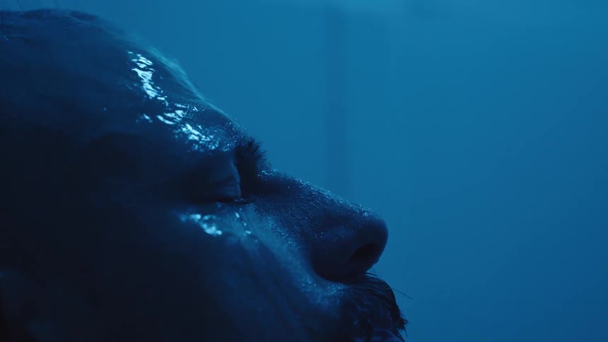 A close side view shows the wet face of a man with shining droplets on his skin and his hand briefly brushing the top of her head