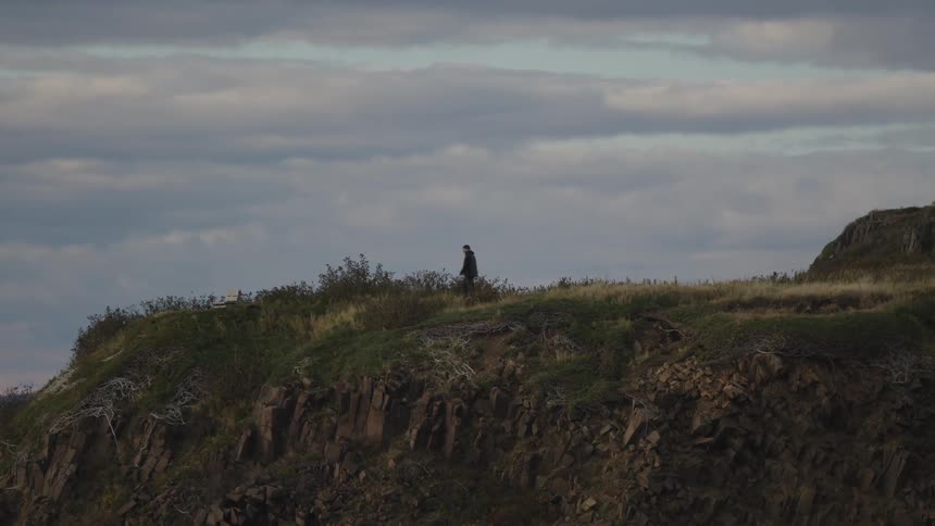 A man walks from left to right across the crest of a rugged rocky cliff covered with grass and low brush against a wide cloudy gray sky