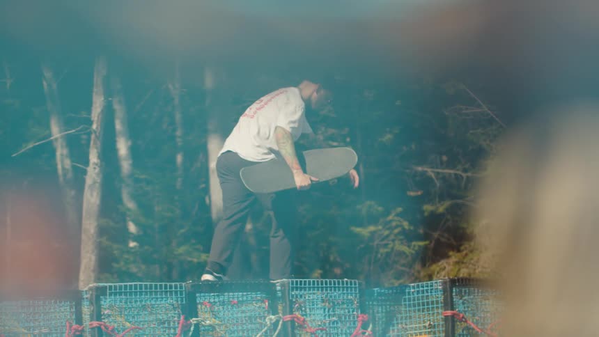 A man walks over a small fence under the midday sun holding a skateboard tucked under his arm as he moves confidently across the open space