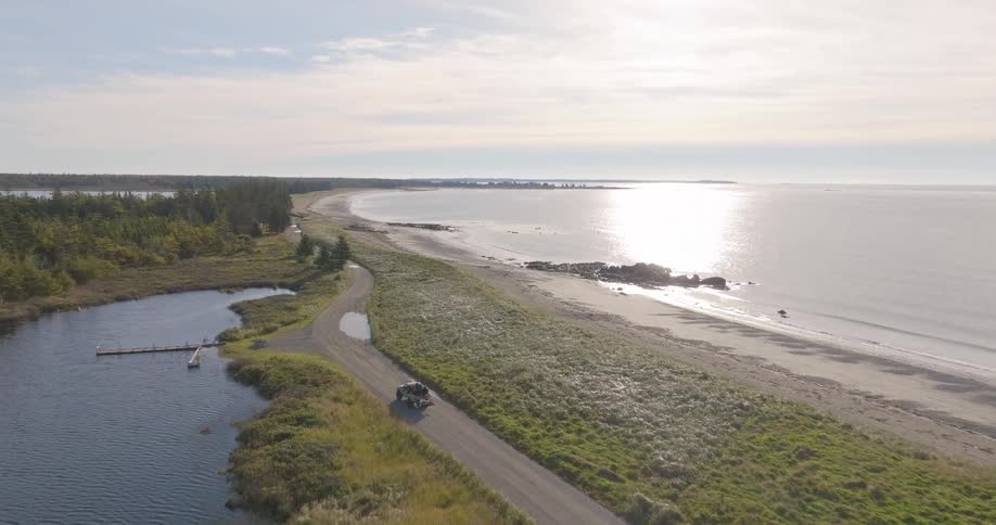 The video is a sweeping aerial view of a dirt road bordering a beautiful coastal area under a bright sunny sky on the left is a dark winding inlet or river and to the right is a wide sandy beach and the sparkling ocean a small offroad